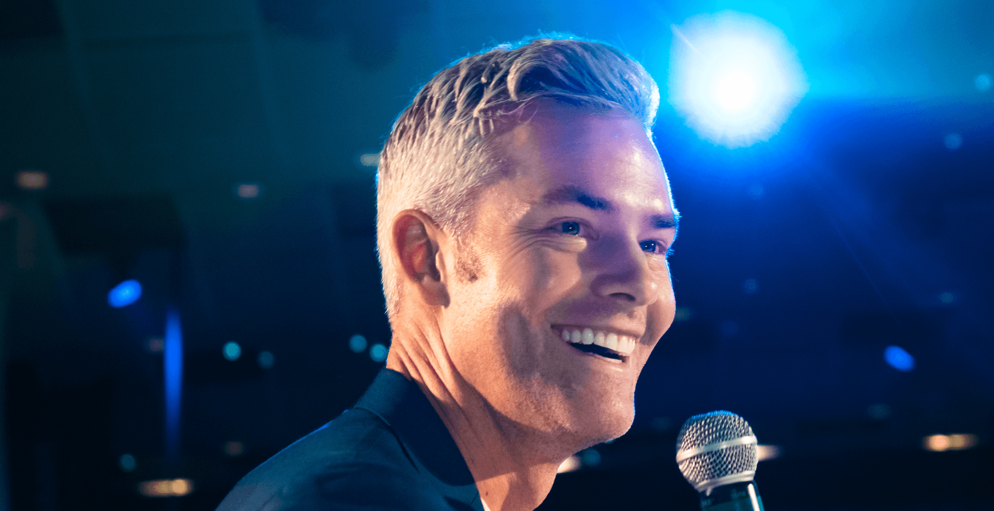 A man with graying hair smiles into a microphone under a blue spotlight.
