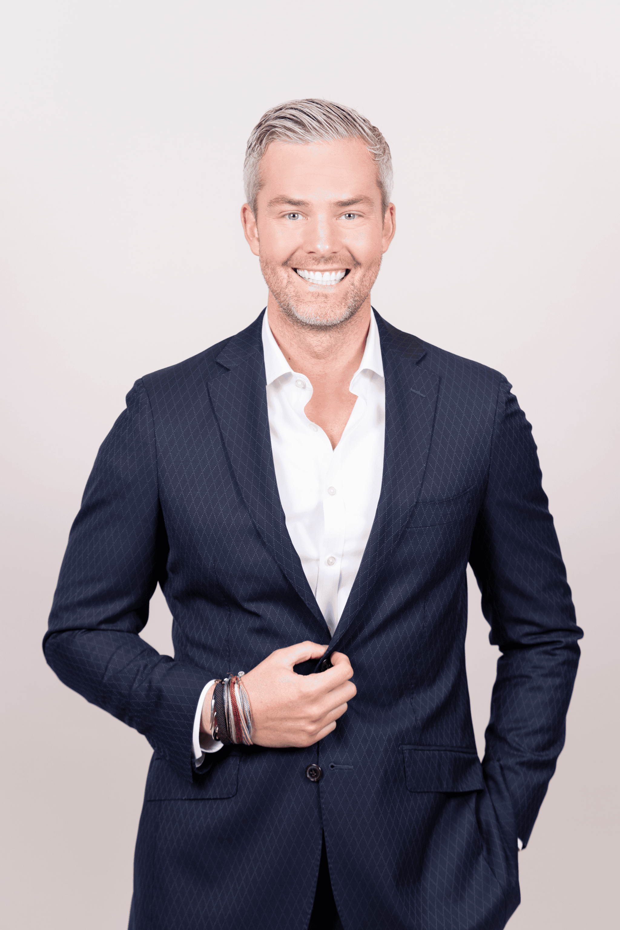 A smiling man with gray hair in a blue patterned suit jacket, holding his lapel, with bracelets on his right wrist.