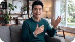 A smiling East Asian man sits on a living room couch, gesturing with his hands.