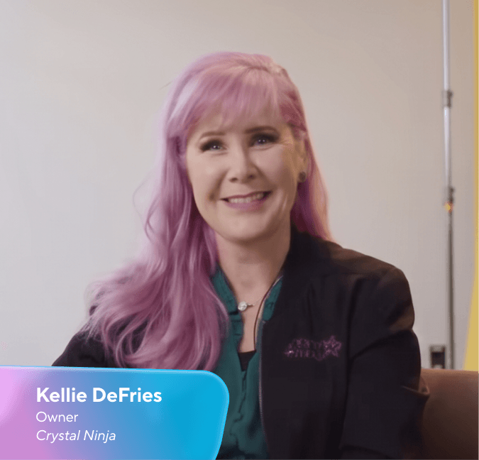 Kellie DeFries, ägare av Crystal Ninja, med rosa hår, leende och klädd i en svart jacka med en lila Crystal Ninja-logotyp.