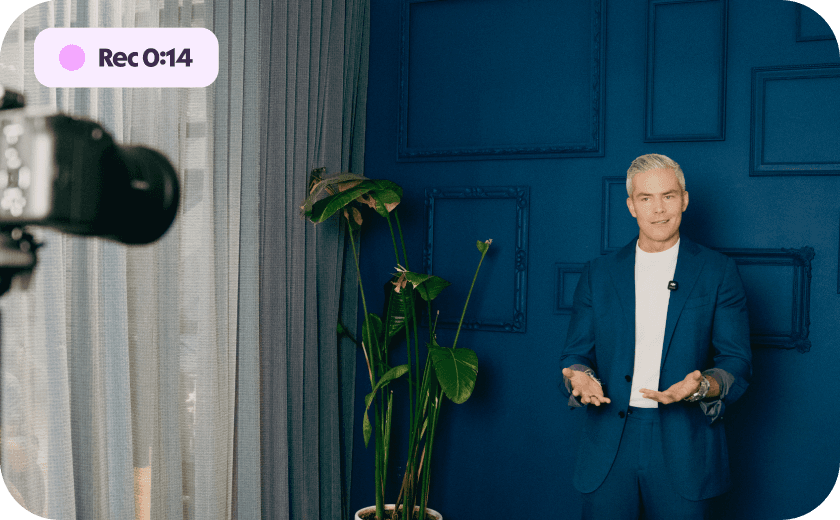 A grey-haired man in a blue suit gestures towards a camera, with a "Rec 0:14" overlay, against a dark blue wall decorated with empty frames and a large plant.
